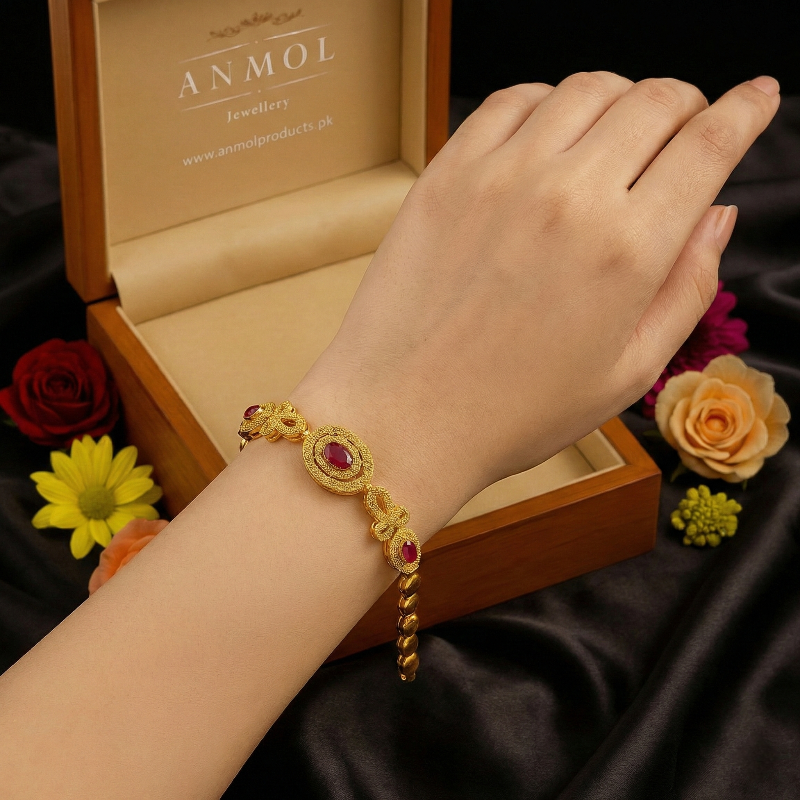 Gold bracelet with red stones on a wrist, against a dark fabric background with flowers and 'ANMOL' jewelry box.
